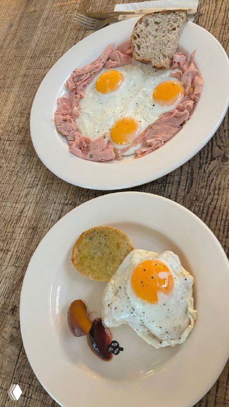 Два блюда на столе: яйца-глазунья и тарелка с гарниром и ломтиком хлеба, на переднем плане видно украшение из мяса и соуса.