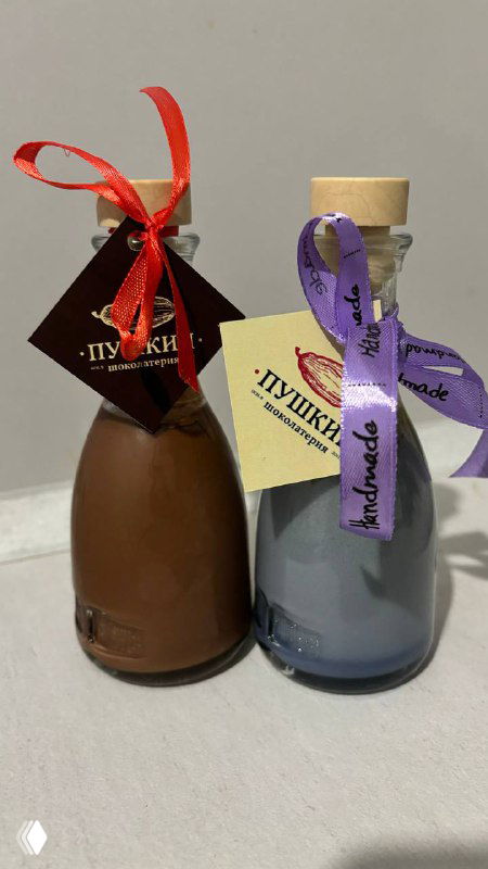 Две маленькие стеклянные бутылочки — шоколадное и лавандовое вино с декоративными ленточками и этикетками, на светлом фоне.