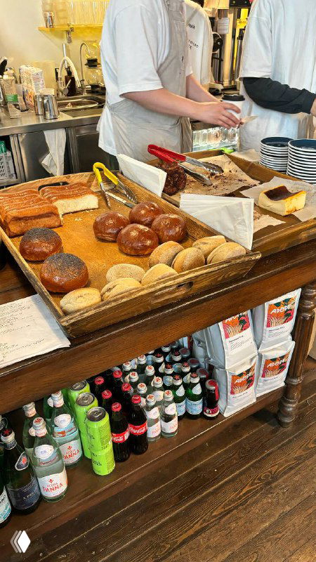 Прилавок с булочками, маффинами и упаковками, видны руки персонала и элементы кассы — типичная сцена в пекарне самообслуживания.