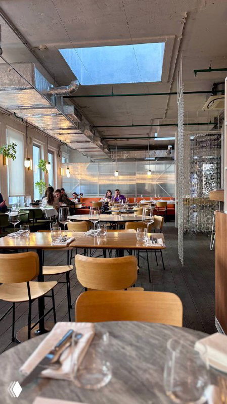 Общий зал ресторана КМ20 с окнами и столами, светлая просторная обстановка и современные деревянные стулья, дневное освещение.