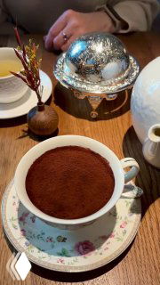 Классический тирамису в холодной тонкой чайной чашке, сверху ровный слой какао; сервировка на столе ресторана Rebecca с серебристой крышкой рядом.