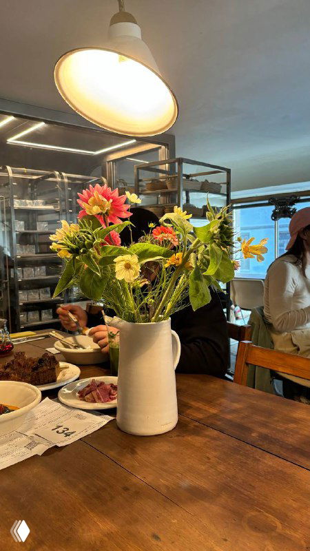 Ваза с цветами стоит на деревянном столе в интерьере пекарни, вокруг гости и спокойная уютная атмосфера заведения.