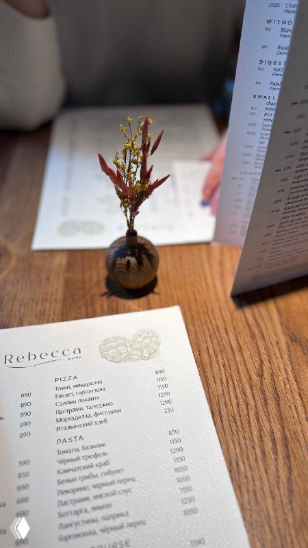 Маленькая керамическая ваза с веткой рядом с меню на деревянном столе — деталь сервировки и эстетики в Rebecca.