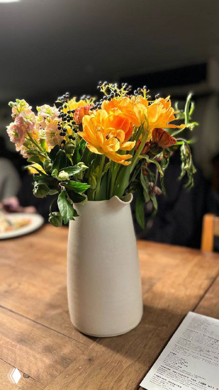 Крупный план другого букета цветов в белой вазе на столе, фон слегка размытый, создаёт атмосферу уюта и внимания к деталям.