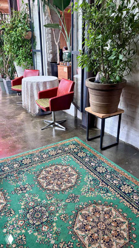 Широкий кадр столика у окна, видны зелёный ковер на полу, горшечное растение, столы и потертая мебель в атмосферном пространстве ресторана.