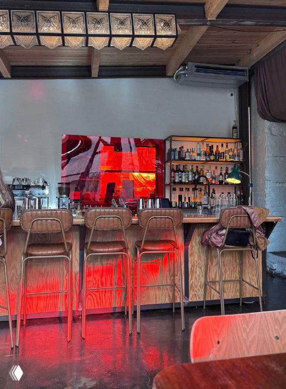 Барная стойка с деревянными стульями и яркой красной подсветкой, пол из шлифованного бетона и стеллаж с бутылками на стене заведения.