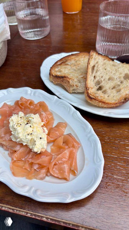 Подача лосося с творожной намазкой и кусочками хлеба на тарелке, видны столовые приборы и ресторанная обстановка.