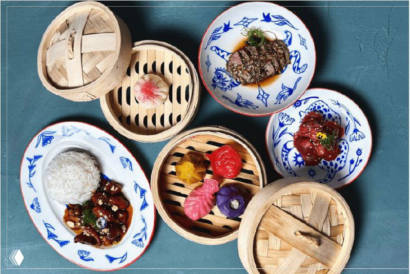 Вид сверху на ресторанную сервировку: несколько тарелок и бамбуковые пароварки с различными закусками и десертами на синем фоне, композиция для гастрономического поста.