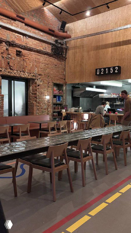 Общий ракурс зала бара с длинным стеклоблоковым столом и рядами стульев, кирпичная отделка стен и открытая кухня на заднем плане.