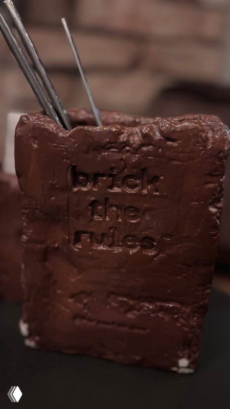 Десерт в форме кирпича с оттиском «brick the rules», крупный план шоколадной глазури и фактуры десерта на тёмном фоне.