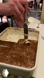 Контейнер с тирамису на столе, официант разрезает десерт лопаткой, видно структуру крема и слоёв какао сверху