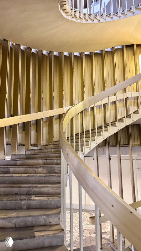 Диагональный ракурс спиральной лестницы: деревянные перила и ступени, повторяющаяся ритмика и мягкая золотистая подсветка в интерьере библиотеки.