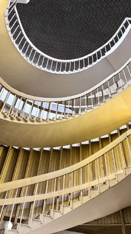 Взгляд снизу вверх по двойной спиральной лестнице: пересекающиеся дуги и геометрические формы, создающие ощущение витка и глубины в библиотеке.