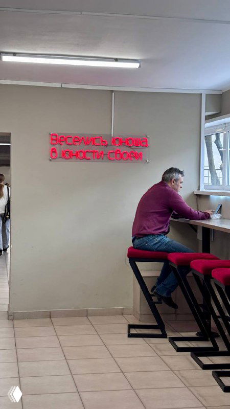 Интерьер столовой или зоны отдыха с неоновым красным текстом на стене и человеком за столом — атмосферный кадр с девизом юности на фоне.