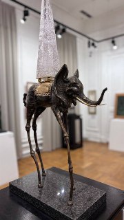 Высокая скульптура слона с тонкими длинными ногами и конусообразным элементом сверху, установлена на черном постаменте в зале.