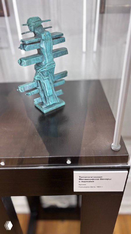 Маленькая бронзовая скульптура современного абстрактного вида в музейной витрине на подставке с подписью экспоната на табличке