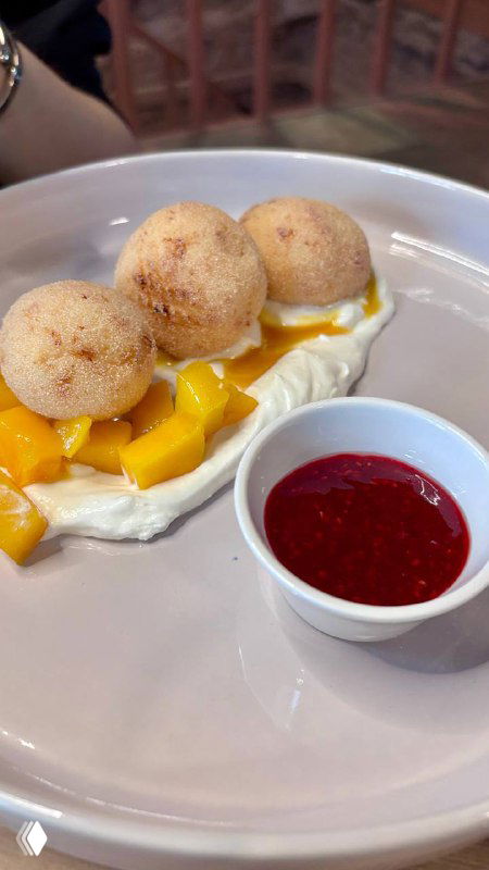 Десерт: шарики мороженого или профитроли на кремовой подложке рядом с кусочками манго и ягодным соусом в маленькой пиале на тарелке.