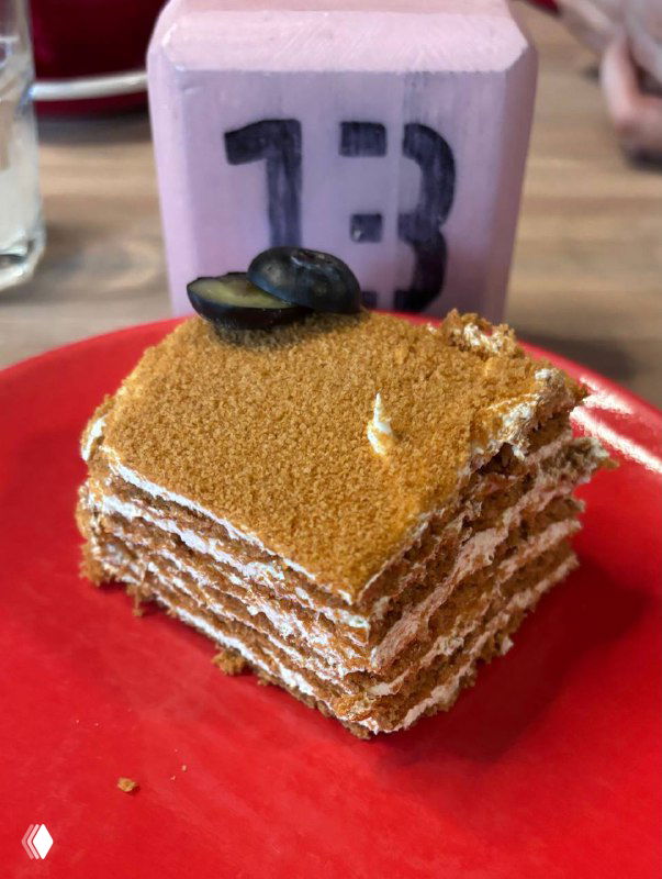 Кусок медовика на красной тарелке: видны слои коржей с кремом и крошка сверху, аккуратно нарезанный десерт в кафе.