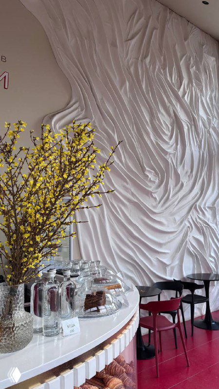 Интерьер кофейни при храме с белой рельефной стеной, веткой желтых цветов в вазе и стойкой с чашками и десертами на переднем плане.