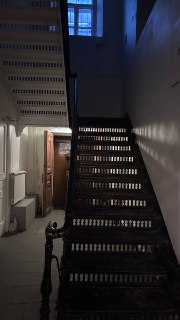 Узкая тёмная лестница в старом здании: деревянные ступени, чёрные перила и длинный коридор с высокими стенами и небольшим окном
