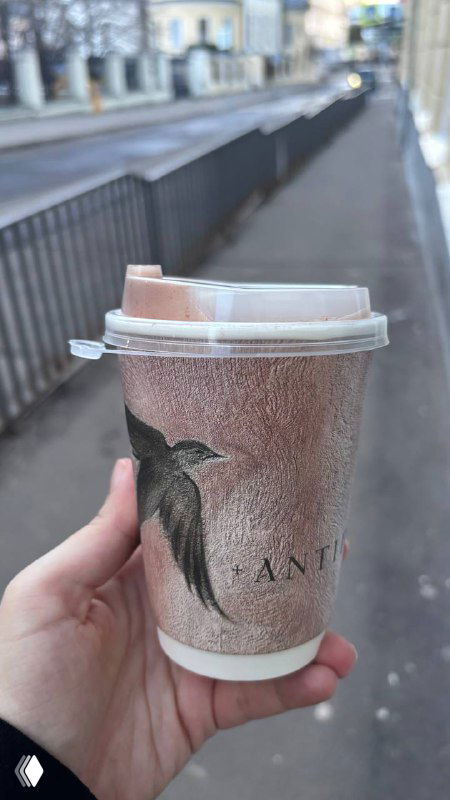 Рука держит бумажный стаканчик с логотипом кофейни на фоне городской улицы — уличный вид и крышка стакана крупным планом.