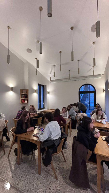 Общий вид зала кофейни при колокольне: высокие потолки, висячие светильники и люди за столиками, атмосфера уюта и света.