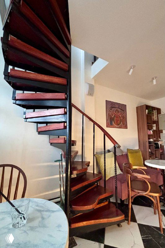 Спиральная деревянная лестница внутри кофейни — вид снизу, ступени и перила образуют виток, интерьер с картинами на стене.