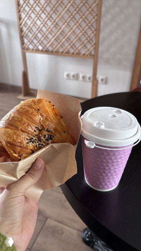 Круассан и бумажный стакан с горячим напитком на столе в кофейне при религиозном здании — завтрак в движении, крупный план.