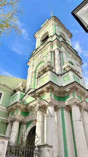 Колокольня с декоративными элементами фасада: столбы, карнизы и высокие ярусы, вид снизу на зелёно-белые поверхности и голубое небо.