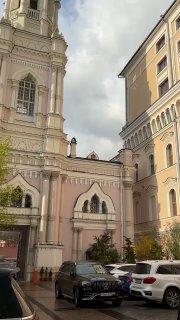Двор храма с фасадом и окружающими зданиями, несколько припаркованных машин, улица и мягкое весеннее освещение в городской среде.
