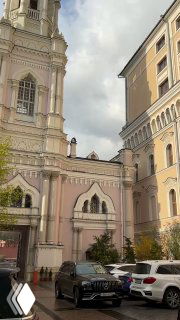 Двор и фасады зданий вокруг храма: пастельные тона стен, припаркованные машины на переднем плане и весенние растения у входа, городской пейзаж.