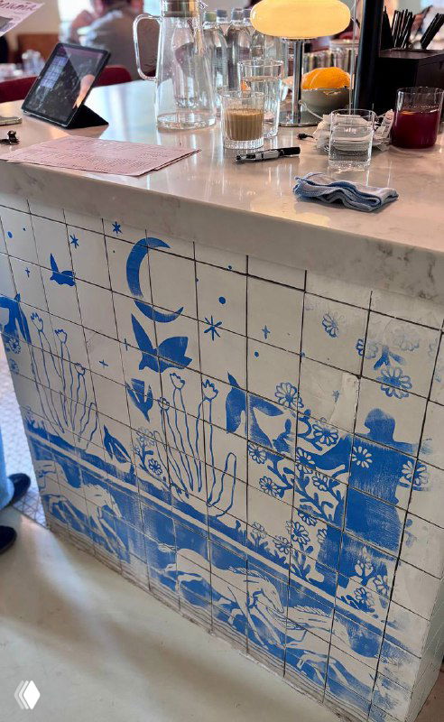 Мраморная стойка бара с керамическими синими плитками, стаканы и бутылки на фоне окна, декоративная посуда на прилавке.