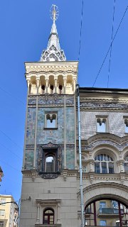 Высокая украшенная башенка и фасад Саввинского подворья на Тверской с мозаичными панно, арочными окнами и ясным синим небом над зданием.