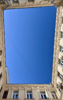 Вид вверх на небосвод через прямоугольный проём двора-колодца, обрамлённый карнизами и окнами четырёх сторон здания на фоне чистого неба.