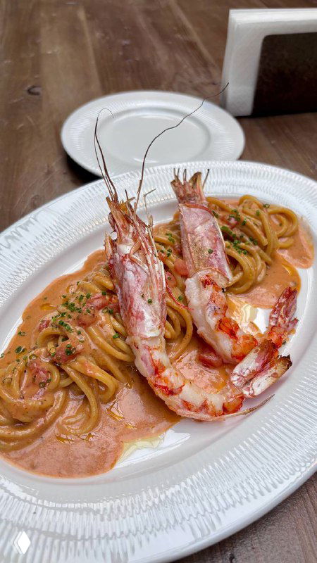 Тарелка со спагетти в кремово-томатном соусе с крупными креветками на белой посуде, снято на деревянном столе внутри светлого кафе.