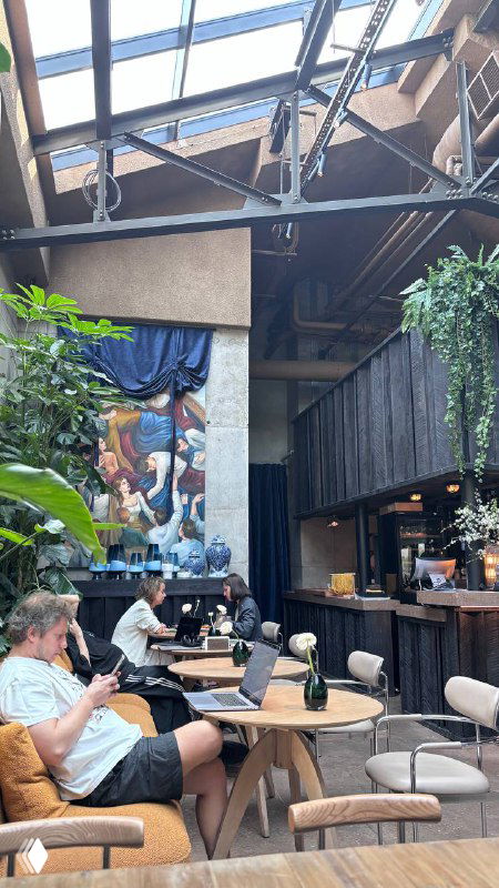 Интерьер кафе с высокими потолками и стеклянной крышей: столы, зелёные растения и гости, ощущение веранды внутри помещения.