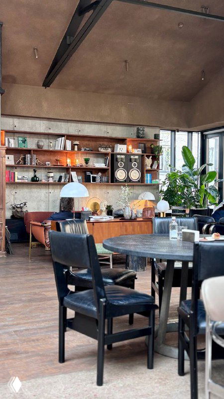 Уютный уголок кафе: столы, стул и открытые полки с посудой и декором, тёплые цвета и много зелёных растений в зоне отдыха.