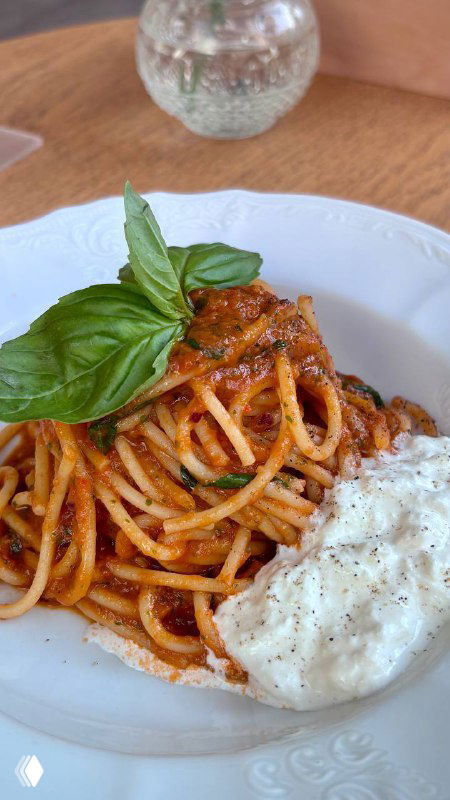 Крупный план спагетти в томатном соусе с кусочком страчателлы и листом базилика на белой тарелке; на фоне деревянный стол и ваза с цветами.