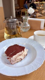 Кусочек тирамису на белой тарелке рядом с прозрачным чайником и чашкой, на фоне деревянный стол и элементы интерьера кафе.