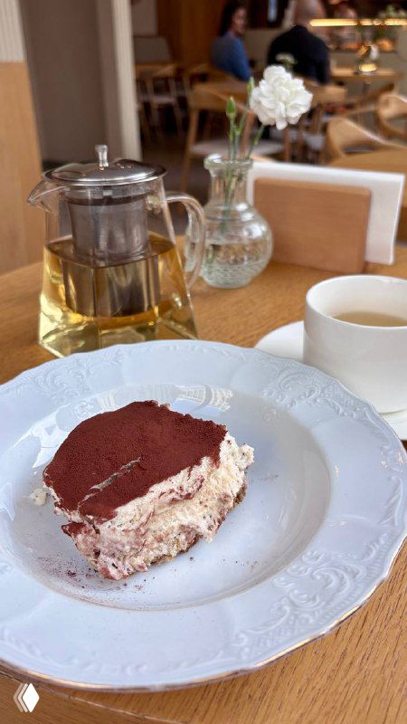 Кусочек тирамису на белой тарелке рядом с чайником и чашкой; десерт снят крупно на столе в уютном интерьере кафе Nobo в Москве.