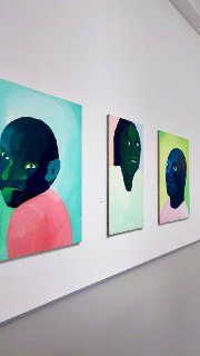 Фото ряда портретных работ: три картины с упрощенными лицами в зеленых и розовых тонах, висящие в один ряд на белой стене галереи.