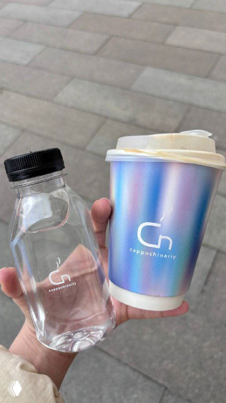 Рука держит фирменный стакан Cappuchinariy с радужным переливом и маленькую пластиковую бутылочку воды на фоне плитки тротуара в Москве