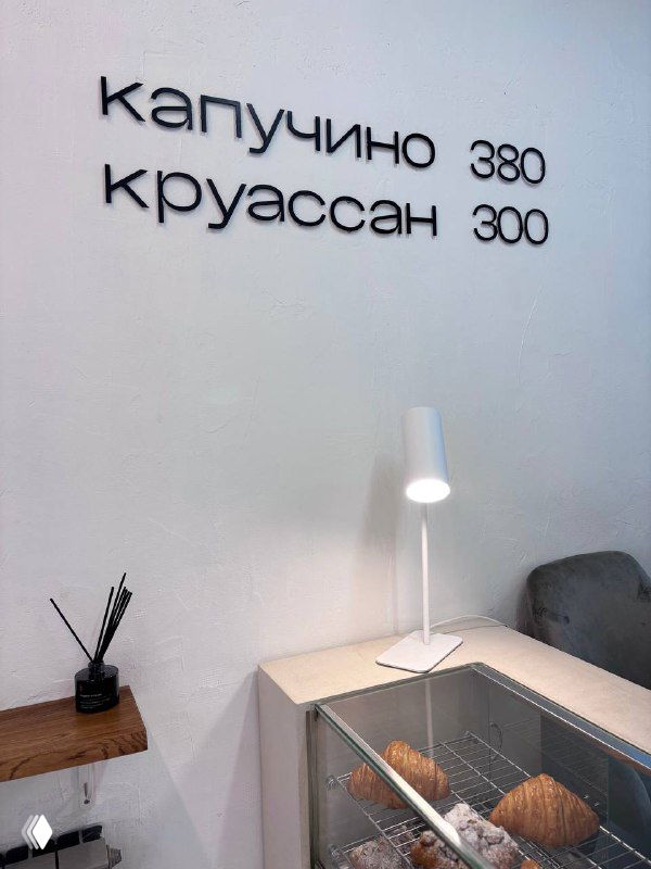 Интерьер кофейни с минимальным меню на стене: надпись «капучино 380 круассан 300», рядом лампа и витрина с выпечкой
