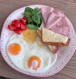 Тарелка с жареными яйцами, ломтиками ветчины, зеленью и помидорами; завтрак подан на узорчатой тарелке крупным планом.