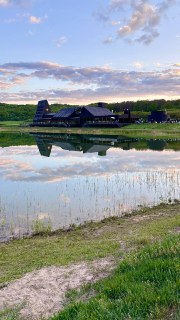 Тёмное современное здание у пруда отражается в спокойной воде; берег с травой и лесной пояс на горизонте
