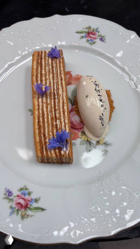 Десерт на фарфоровой тарелке: медовик в дуэте с шариком подсоленного мороженого, украшены съедобными синими цветами, крупный план.