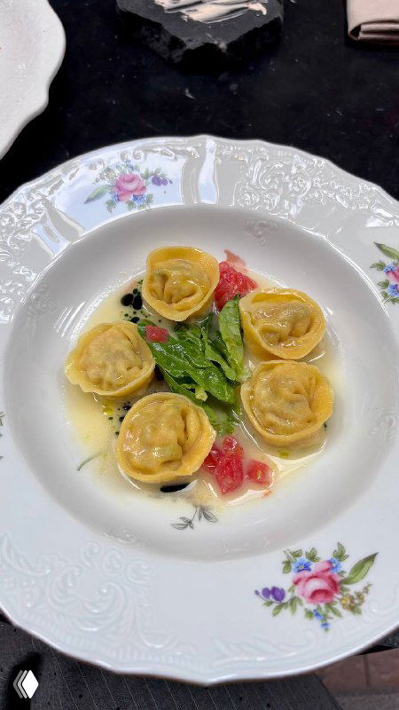 Тарелка с равиоли в светлом соусе, зелень и кусочки томата на фарфоровой посуде с цветочным узором, вид сверху.