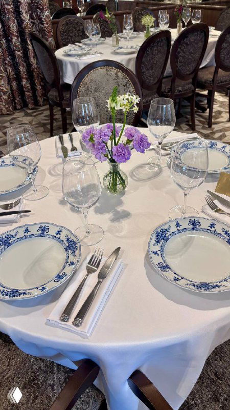 Круглый стол с белой скатертью, гжельские тарелки и стеклянные бокалы; в центре ваза с фиолетовыми и белыми цветами — деталь сервировки.