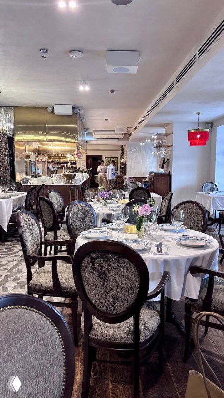 Ряд столов в ресторане с круглыми стульями и аккуратной сервировкой, в глубине видно открытую кухню и светлые потолки.