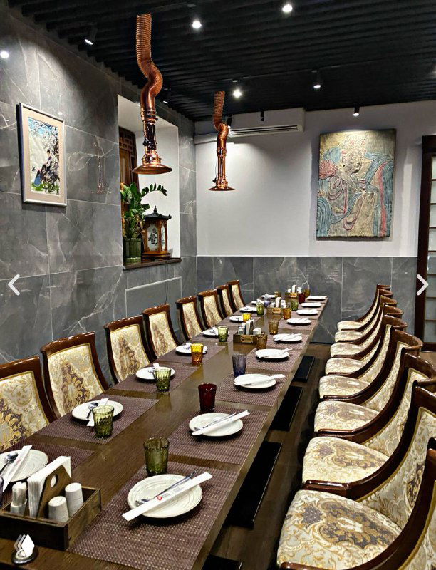Интерьер корейского ресторана в Москве: длинный общий стол для компании, резные стулья, настенные картины и медные вытяжки над столом.